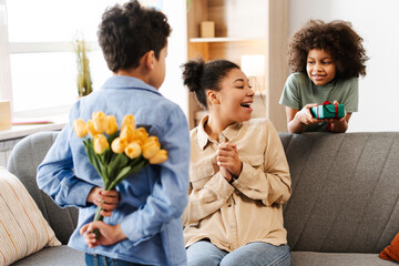 African American mother, receiving presents from her kids. Son conceals bouquet, ready to surprise. Mother's Day concept  - obrazy, fototapety, plakaty