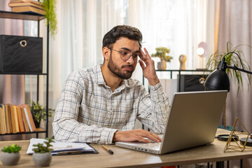 Bored sleepy young businessman worker working on laptop computer, leaning on hand at home office desk table. Exhausted tired Arabian freelancer workaholic guy. Employment, occupation, workless.