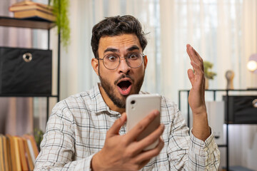 Oh my God, wow. Happy amazed young Hispanic businessman working on smartphone shocked by sudden...