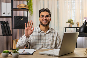 Hello. Happy young Indian businessman employee waves hand hi gesture greeting welcomes someone, online webinar, remote distance conversation. Freelancer guy sitting at desk with laptop in home office.