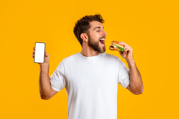 man enjoys cheeseburger while showcasing blank mobile phone screen, studio