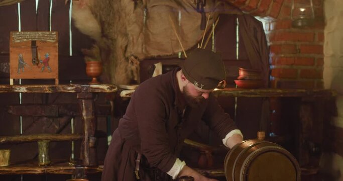 Medieval keeper pours local wine from barrel at inn. Barkeeper in ancient clothes fills clay mug with alcohol in tavern. Concentrated servant in pub
