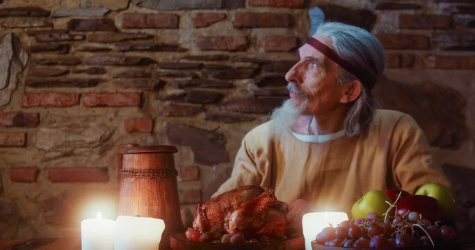 Old man drinks and looks aside in tavern. Elderly aristocrat has abundant banquet with grilled chicken and alcohol in castle. Rich medieval lord dinner