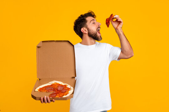 guy holding delivery box and savoring pizza slice, yellow backdrop