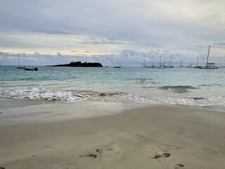 captivating scene in Guadeloupe with a sandy beach bordered by a distant islet and sailboats on the horizon. Waves dance under overcast skies, creating a mysterious ambiance in this tropical enclave
