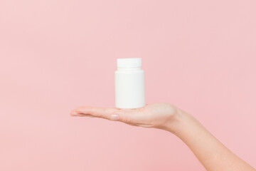 White bottle plastic tube in woman's hands on pink background. Packaging for pills, capsules or supplements. Cosmetics
