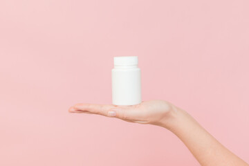 White bottle plastic tube in woman's hands on pink background. Packaging for pills, capsules or...