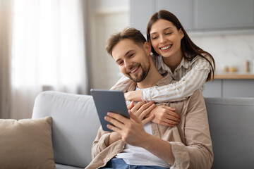 Happy woman embracing husband while using tablet on sofa