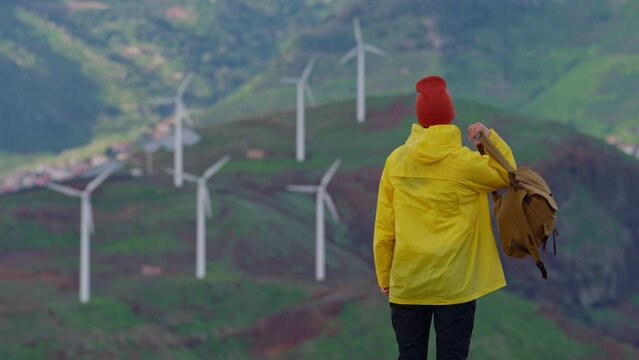 Attractive traveling woman in a yellow jacket and backpack looking at windmills in field 
