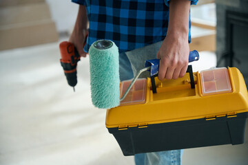 Man hands holding electric screwdriver, paint roller and tool box