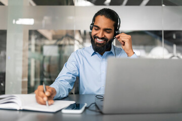 Cheerful Indian man with headset working online on laptop computer