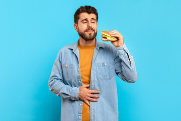 Young man touches his aching stomach holding burger in studio