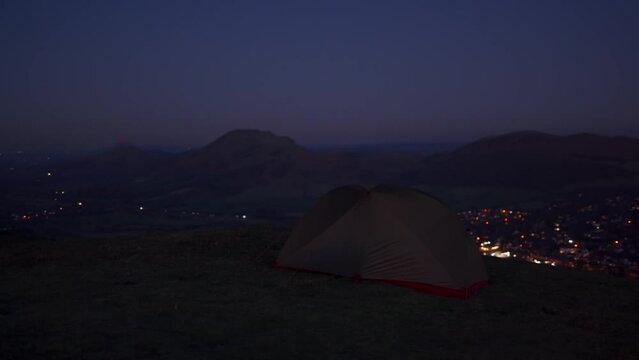 A man standing beside his wild camping tent on a hill overlooking a town and rural British scene