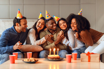 Diverse friends embracing birthday guy sitting with party hats indoors