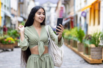 Happy Indian young woman standing on city street in green suit and looking at phone screen, happy to receive news and message showing victory gesture with hand