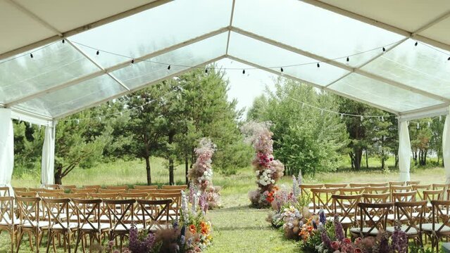 Outdoor wedding ceremony under the tent in garden among the trees. View of beautiful and tender wedding floral arch and wooden chairs on green lawn slow motion. 