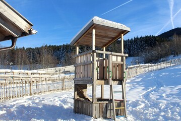 Kletterturm aus Holz für Kinder auf einer Skipiste. Im Hintergrund eine Übungspiste, hölzerner Zaun. Natur, Winterlandschaft, Allgäu, Bayern.