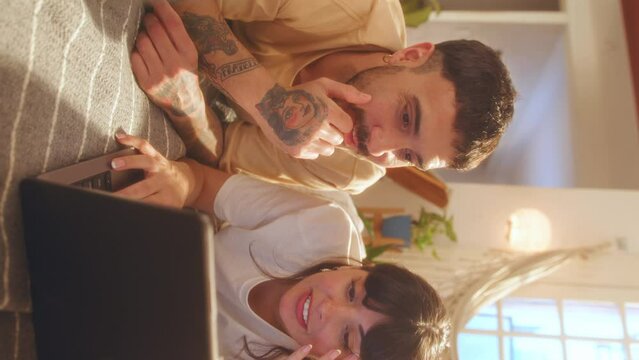 Young happy boyfriend and girlfriend lying on bed and discussing something on a laptop screen, surfing the Internet or doing online shopping during leisure time at home. Vertical format footage