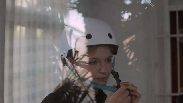 A beautiful blonde puts a white bicycle helmet on her head. The video was shot through the window. Concept of generation alpha, generation Z, diversity.