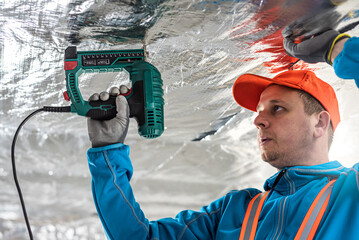 The builder is fastening the vapor barrier under the ceiling thermal insulation using an electric...