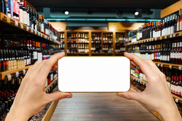 Unrecognisable smart phone with blank screen in hands in front of shelves with wine bottles in a...
