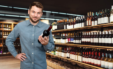 Man customer holding wine bottle in hand and smiling in liquor store.