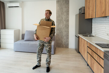 Smiling man in camouflage holding cardboard box in new apartments