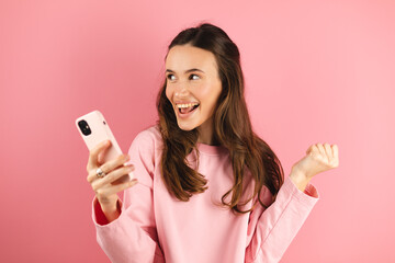 Amazed brunette woman look surprised wow hold using mobile cell phone on pink background. Girl look...