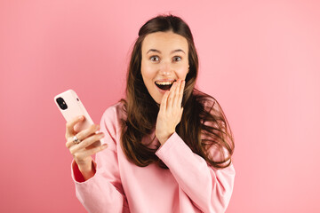 Shocked brunette woman wear pink long sleeves top hold mobile phone isolated on pink background...