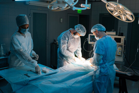 Skilled Cooperating Team Of Doctors And Surgeons Processing Surgical Operation In Dark Operating Room Modern Hospital Emergency Department. Doctors Wearing Sterile Gloves, Surgical Uniforms And Masks.