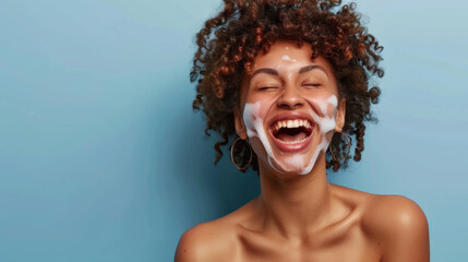 A cheerful woman with a skincare cream applied on her face is laughing with eyes closed against a blue background.
