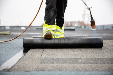 professional roofer applying tar with blowtorch on new roofing felt for waterproofing a flat roof construction site with safety gear - obrazy, fototapety, plakaty