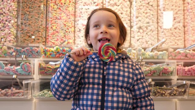 In a sweets store girl child cheerfully licks a large lollipop