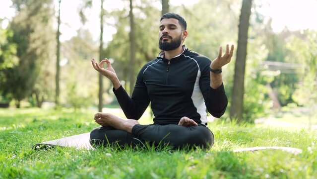 Adult bearded man meditating in the lotus position on a mat in an urban city park. Handsome male in sports suit doing yoga outside. Concentrated man practices meditation barefoot with his eyes closed