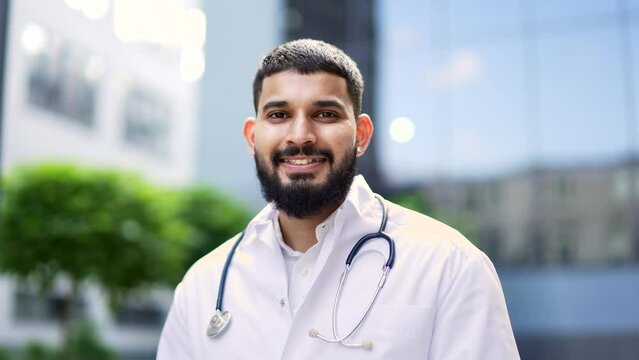 Portrait of a confident smiling bearded doctor in a white coat standing outside near a hospital building. Happy handsome medical worker physician with stethoscope posing looking at camera. Close up