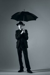 a full-length portrait of a young man in a suit with a hat and an umbrella, shot in a studio in black and white