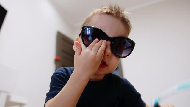 Funny Caucasian baby boy wearing big black sunglasses. Portrait of a cute kid at home. Close up.