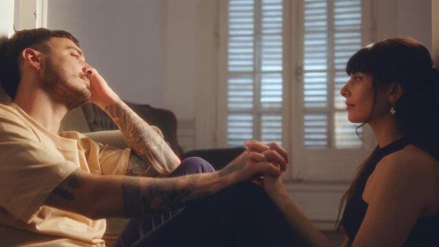 Young Couple In Love Sitting On The Floor In Doorway, Holding Hands And Eye Contact During Romantic Evening At Home. Side View