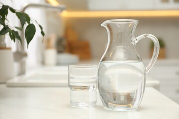 Jug and glass with clear water on white table in kitchen, space for text