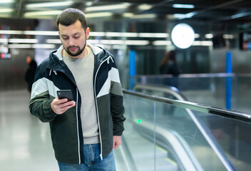 Portrait of positive bearded guy traveler standing on empty subway station with smartphone in...