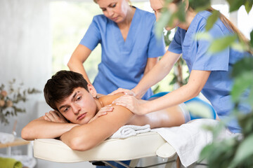 Two female masseur doing massage at back man who lying on massage table in a beauty salon