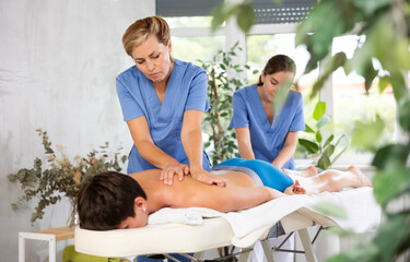 Two female masseur doing massage at back man who lying on massage table in a beauty salon