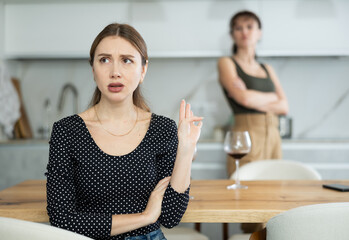Sad middle-aged woman sitting at table and other woman brawling to her behind in the kitchen