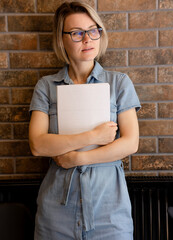 Blonde student girl in glasses and blue dress holding laptop in her hands. Woman standing near brick wall and clutching gray laptop for work, interview, freelancing, student