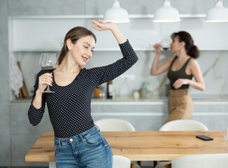 Woman has drunk wine and is dancing merrily to music in kitchen. In background, friend with glass...