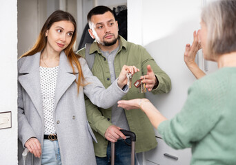 Portrait of young positive family couple standing at doorway and giving keys to mature woman owner...