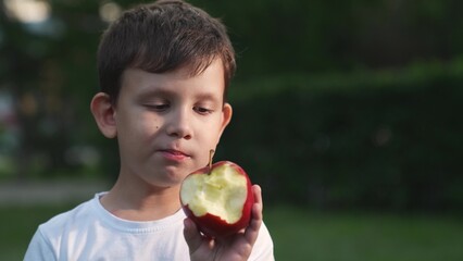 Funny little boy eating fresh appetizing red apple enjoy taste at summer park outdoor closeup. Adorable male kid child bite raw organic vitamin fruit chewing delicious edible plant healthy lifestyle - obrazy, fototapety, plakaty