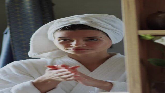 Vertical close-up of Caucasian or Hispanic woman in bathrobe, wet hair in towel turban, taking dollop of lotion from bottle, rubbing between palms, spreading over face while looking in mirror