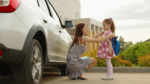 Young caring mother help little daughter putting on backpack going to primary school outdoor slowmo. Happy family woman and girl kid child getting ready to classroom elementary lesson at schoolyard