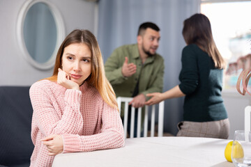 Stressed young woman sitting at table while family couple of her friends arguing behind